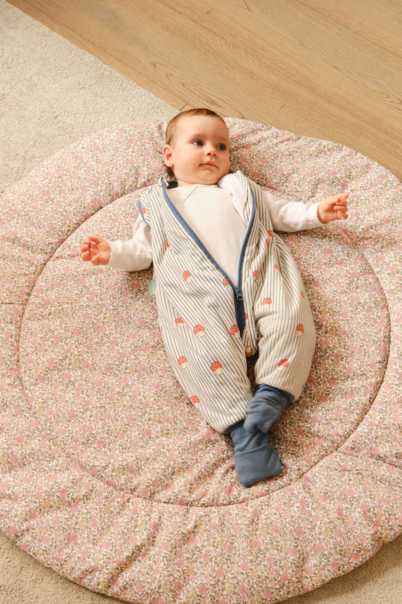 Bebé deitado em almofada floral com babygrow branca e saco de dormir às riscas com cogumelos