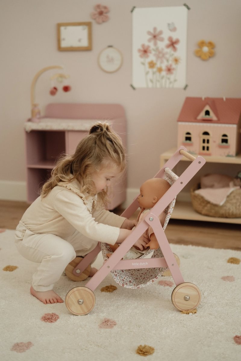 Menina a brincar com carrinho de boneca rosa em madeira e tecido flores num quarto com tapete e móveis pastel