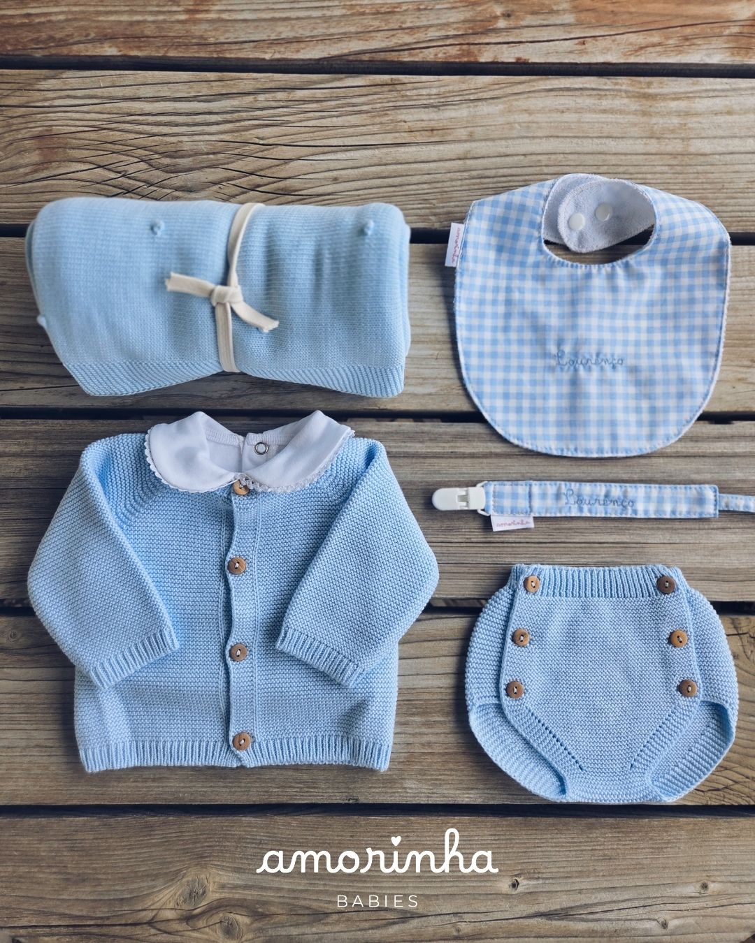 Conjunto de roupa de bebé azul com casaco, fralda, babete e faixa para chupeta sobre fundo de madeira