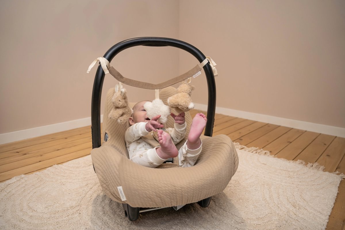 Cadeira de bebé bege com bebé e brinquedos de peluche, em ambiente interior com chão de madeira e tapete claro.