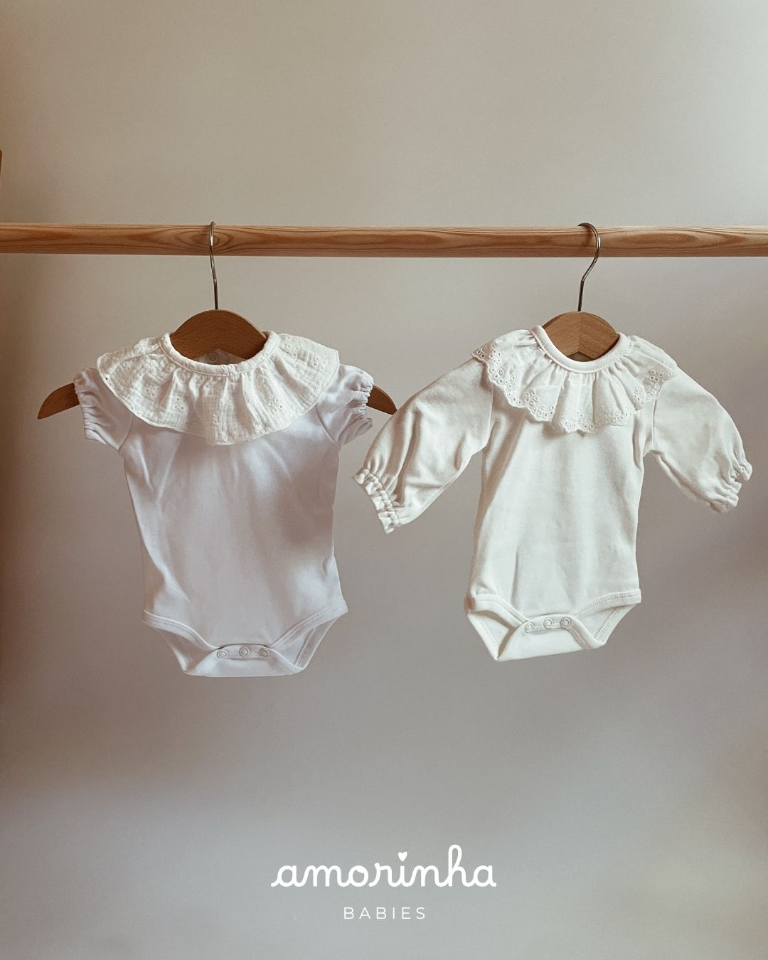 Duas roupas de bebé brancas penduradas em cabides numa barra de madeira