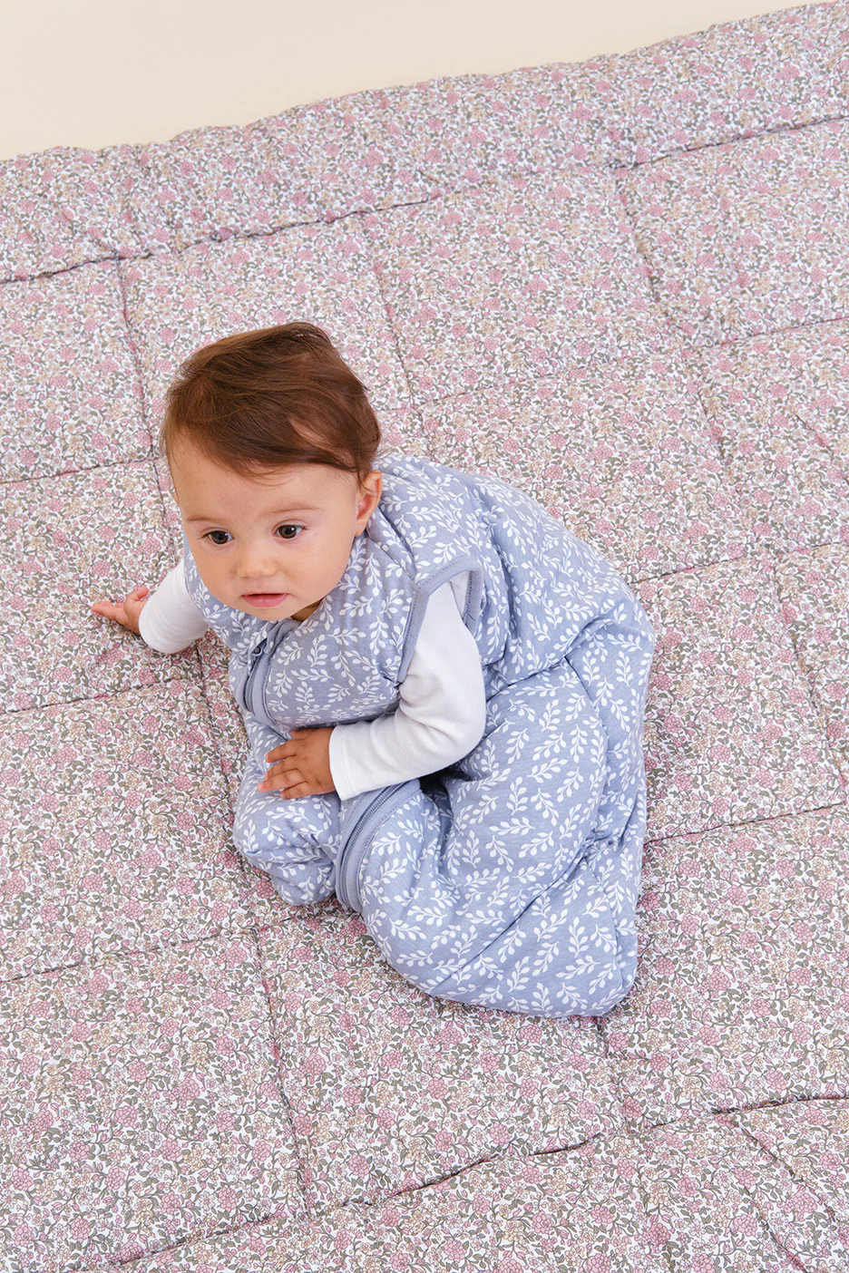 Bebé em saco de dormir azul com padrão de folhas sentado em tapete floral