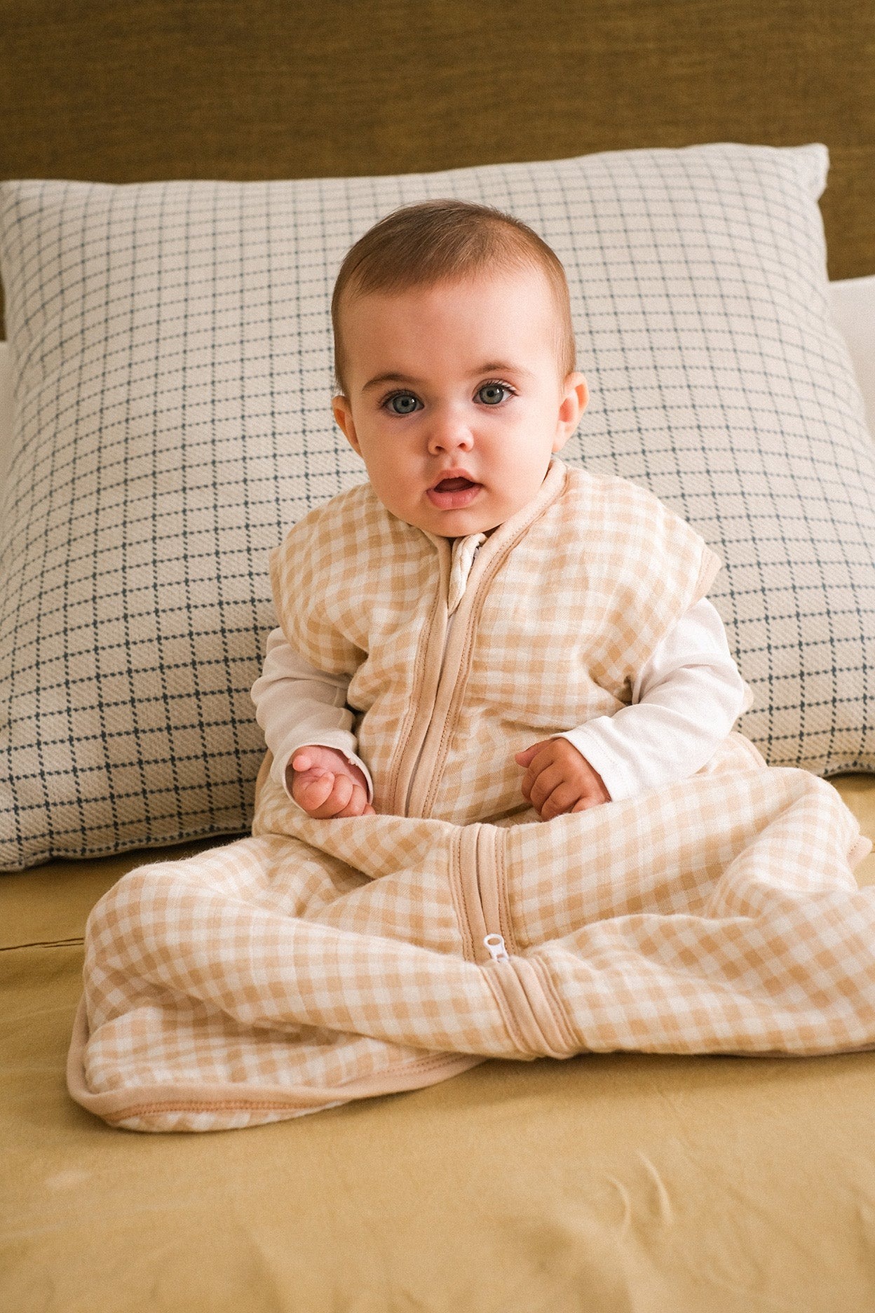 Bebé com saco de dormir bege claro aos quadrados sentado numa cama com lençol amarelo e almofada xadrez azul e branca.