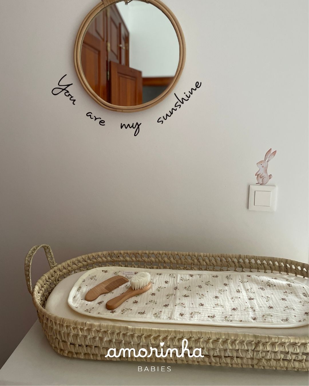 Cesta de bebé de vime com toalha floral e acessórios de madeira sob espelho redondo na parede com texto decorativo