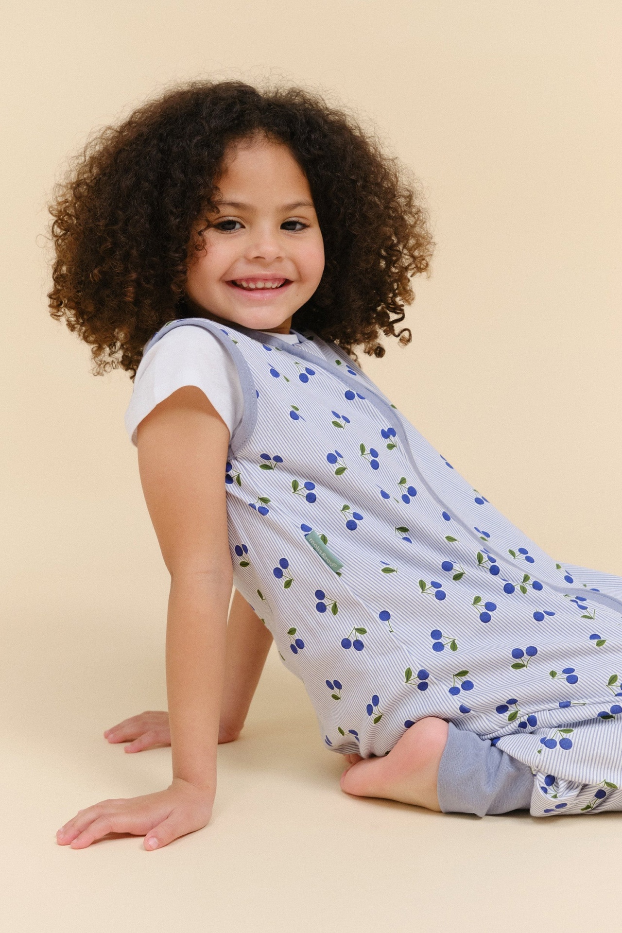 Menina com pijama azul claro de cerejas sentada em fundo bege.