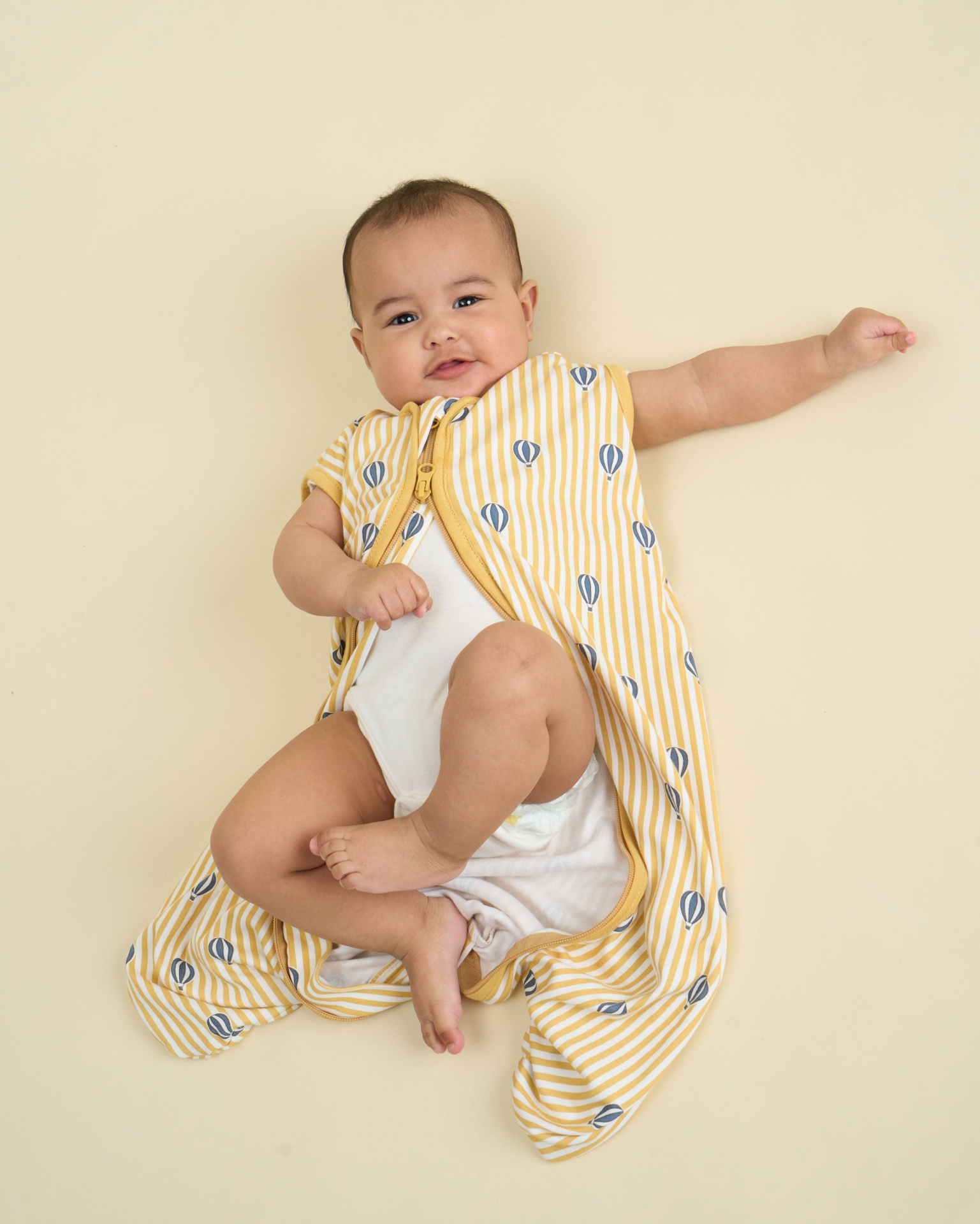 Bebé com saco de dormir amarelo e body branco a sorrir sobre fundo amarelo claro