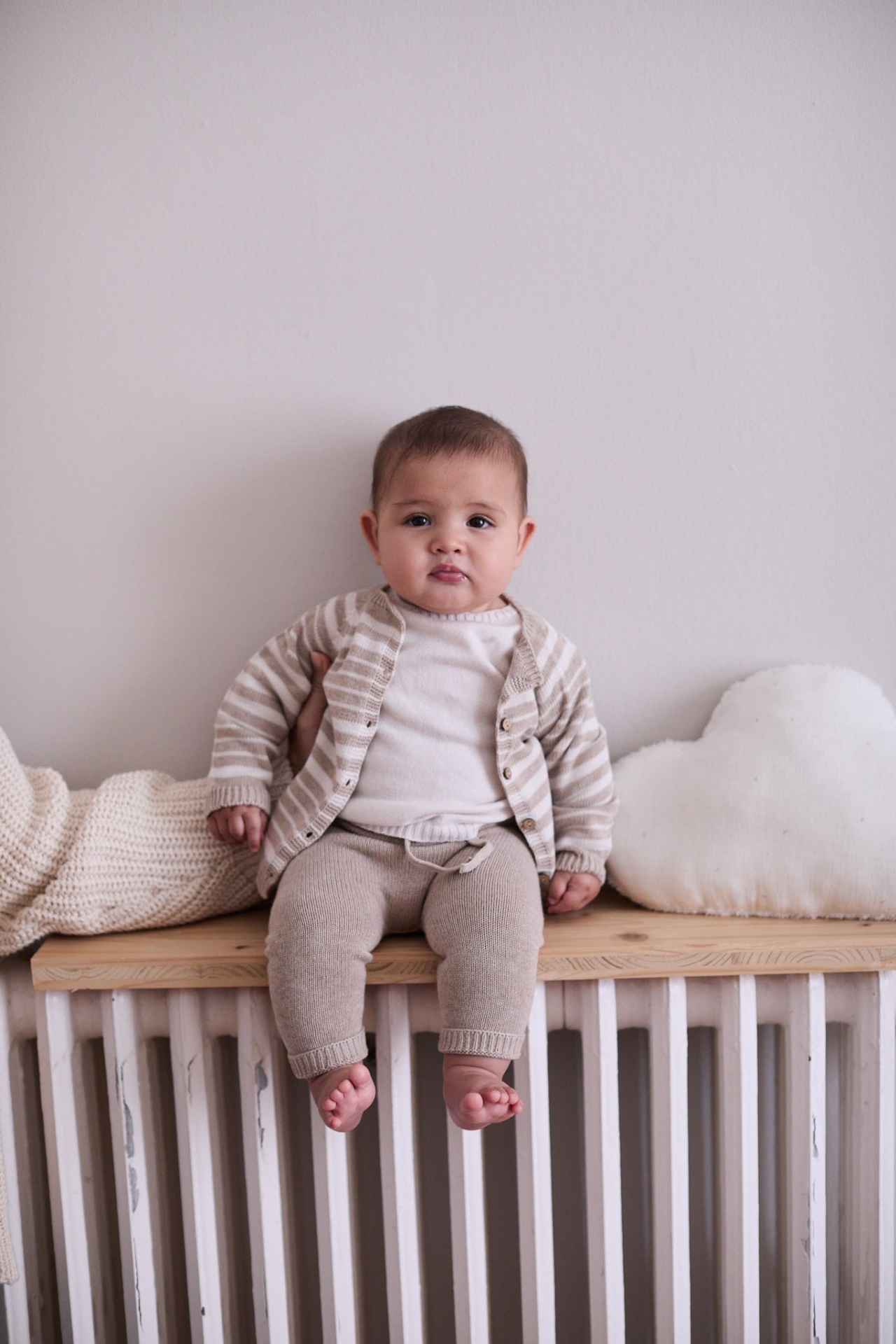 Bebé com roupa de malha bege e creme sentado num banco de madeira com almofada em forma de nuvem