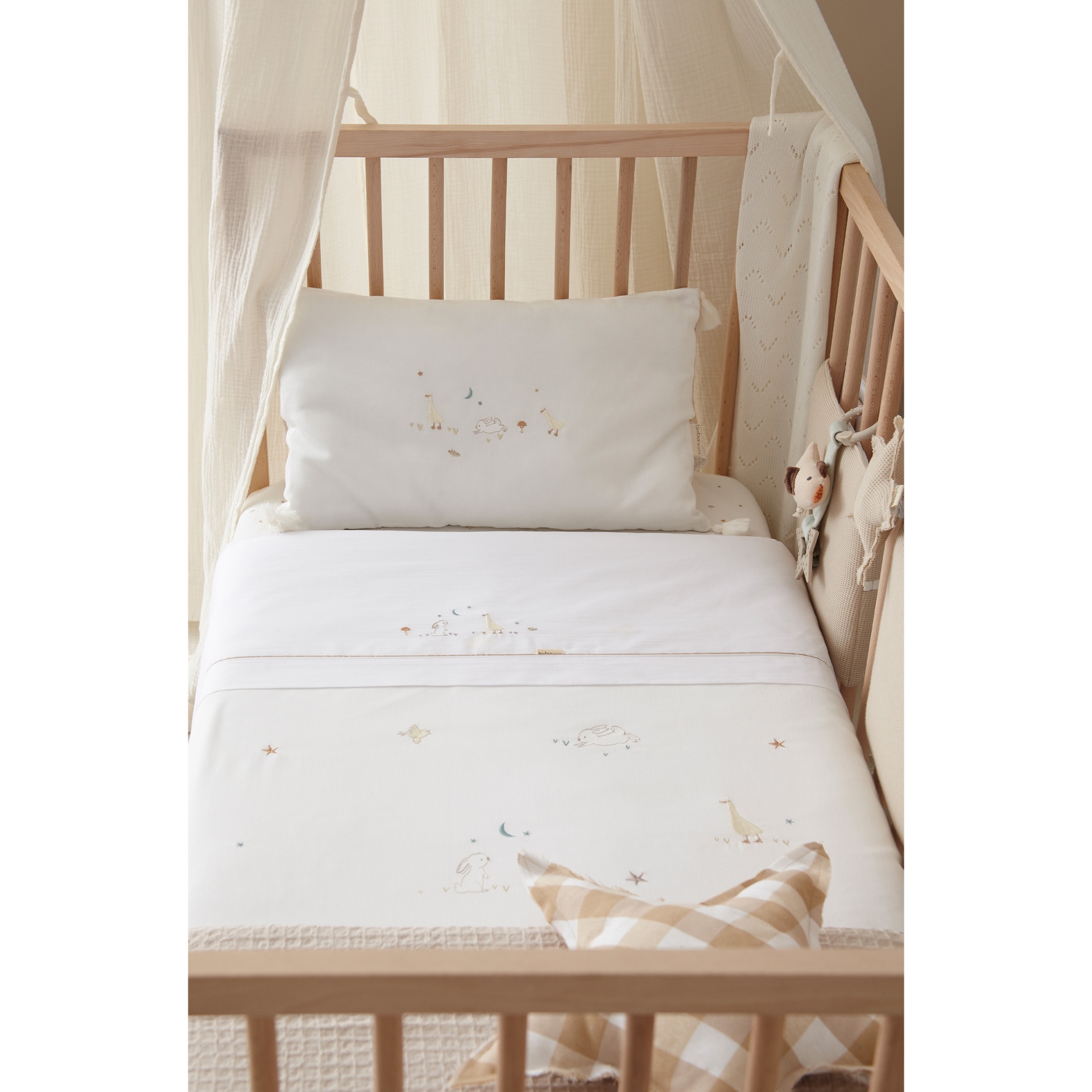 Berço de madeira clara com tecido leve bege e roupa de cama branca com bordados infantis