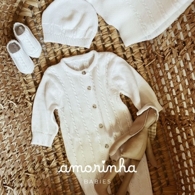 Conjunto de roupa de bebé branco com macacão, gorro e botinhas sobre tapete de fibras naturais.