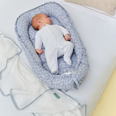Bebé deitado num berço de tecido azul com padrão branco, macacão branco e manta branca com borda cinzenta num colchão branco