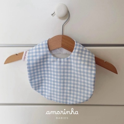 Babete de bebé azul e branco com padrão quadriculado pendurado num cabide de madeira.