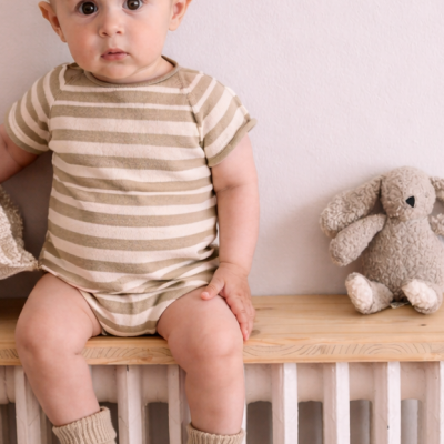 Bebé com body às riscas bege e branco e meias bege sentado ao lado de um coelho de peluche numa superfície de madeira.