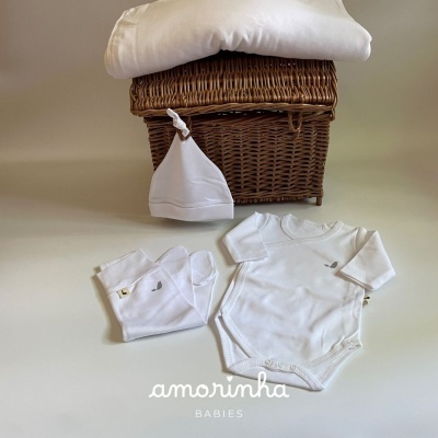 Conjunto de roupa de bebé branca com body, calças e gorro em frente a um cesto de vime e cobertor branco