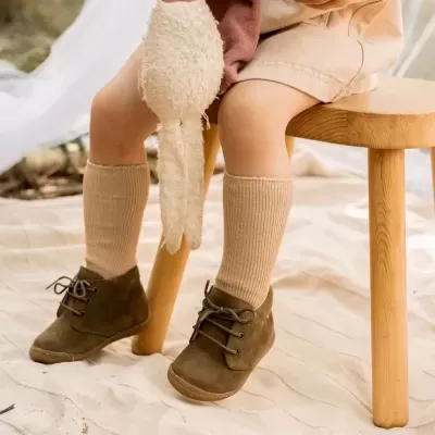 Criança com meias castanhas claras e sapatos castanhos segurando brinquedo de tecido sentada em banco de madeira