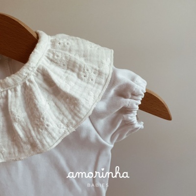Blusa branca para bebé com gola rodada texturizada com padrão floral bordado pendurada num cabide de madeira