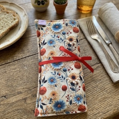 Estojo de tecido floral amarrado com fita vermelha numa mesa de madeira com talheres e copo.