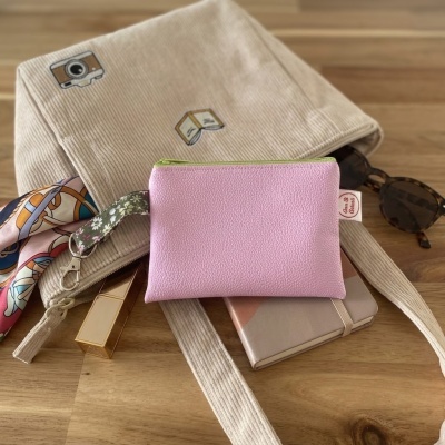 Bolsa pequena rosa com alça floral sobre mochila bege com bordados, óculos, batom e caderno sobre mesa de madeira