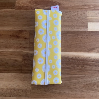 Estojo amarelo com margaridas brancas e fecho elástico branco sobre madeira.