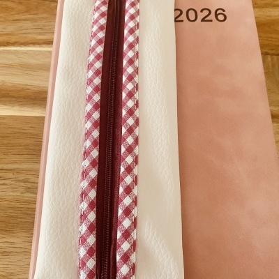 Estojo branco com zipper vermelho e branco xadrez sobre agenda rosa 2026 em mesa de madeira