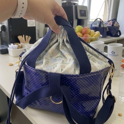 Saco azul com pontos brancos e interior floral, segurado por mão com pulseira branca, sobre mesa com utensílios.