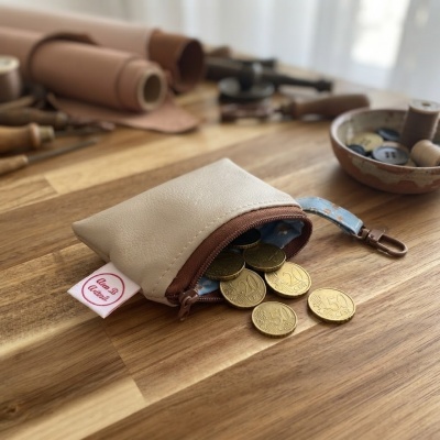 Carteira pequena bege em pele artificial com fecho castanho e moedas douradas a sair, em mesa de madeira com ferramentas e botões.