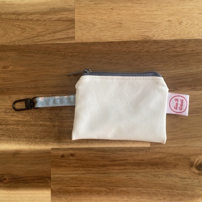 Bolsa pequena branca com fecho cinzento e alça azul estampada com mosquetão preto sobre fundo de madeira.
