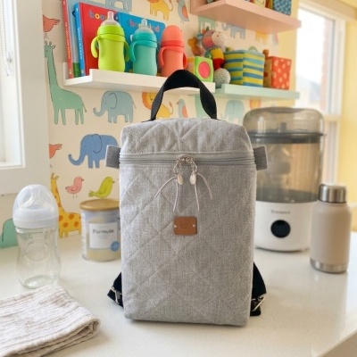 Mochila térmica cinza acolchoada com alça preta numa cozinha com acessórios de bebé e papel de parede com animais.