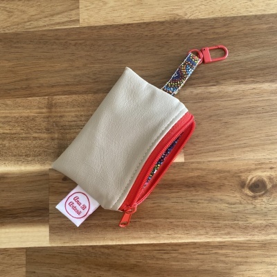 Bolsa pequena bege com fecho vermelho e cinta colorida com padrão de bolinhas sobre superfície de madeira