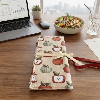 Guardanapo de tecido com padrão de maçãs e fita decorativa numa mesa de trabalho com laptop, planta, salada e copo.