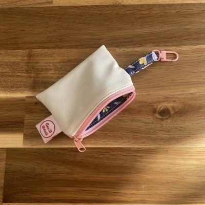 Bolsa pequena branca com fecho rosa e mola cor de rosa, com fita azul floral, sobre superfície de madeira.