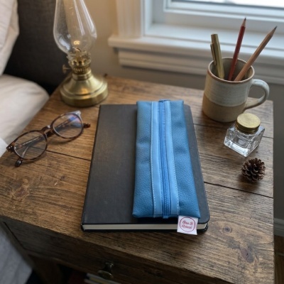 Mesa de cabeceira em madeira com lâmpada, óculos, caderno, estojo azul, caneca com lápis, frasco de vidro e pinha
