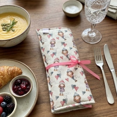 Guardanapo de tecido estampado com desenhos de meninas e corações, amarrado com fita rosa sobre mesa com talheres e comida