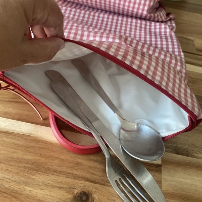 Bolsa de tecido xadrez vermelho com talheres metálicos dentro aberta sobre mesa de madeira