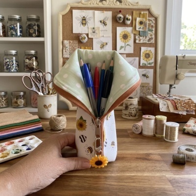 Estojo branco com padrão de abelhas e fecho com pendente de girassol aberto e cheio de canetas, sobre mesa de madeira com material de costura ao fundo.