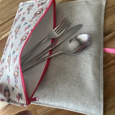 Estojo organizador de talheres em tecido com padrão bonecas e fecho rosa aberto com talheres de metal.