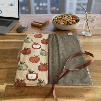 Toalhete de tecido com padrão de maçãs e fita castanha sobre mesa de madeira com salada, copo com água, rádio e computador ao fundo.