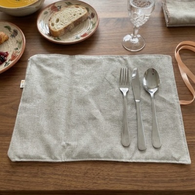 Talheres metálicos sobre bolso de tecido cinza claro em tabuleiro de madeira com pratos decorados e sopa ao fundo.
