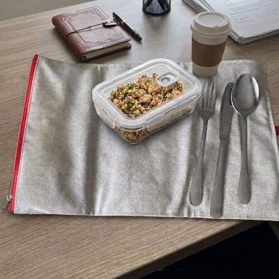 Talheres em almofada cinza, recipiente plástico com comida, copo descartável, caderno e canetas em secretária