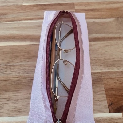 Estojo rosa com fecho bordô contendo óculos dourados e caneta, sobre madeira clara