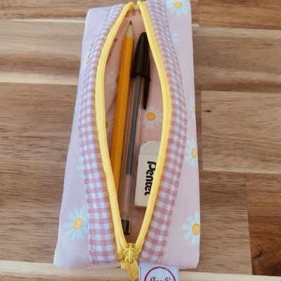 Estojo de tecido com padrão rosa e margaridas, fecho amarelo, contendo lápis, caneta e borracha sobre madeira.