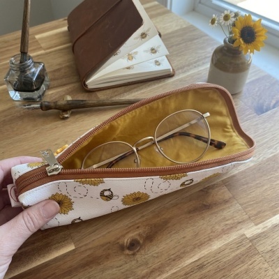 Estojo para óculos branco com girassóis e abelhas, interior amarelo, com óculos dentro, sobre mesa de madeira com caderno e tinteiro ao fundo