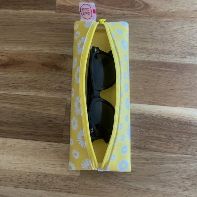 Estojo amarelo com margaridas que contém óculos de sol pretos Ray-Ban sobre madeira.