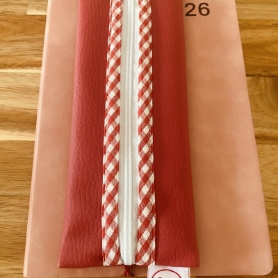 Bolsa vermelha com fecho branco e padrão xadrez, etiqueta Ana B Artesã, sobre superfície de madeira e objeto rosa com número 26.