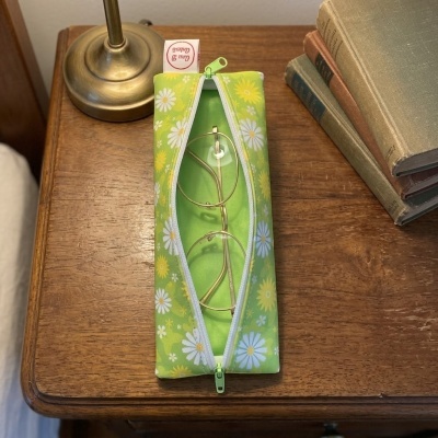Estojo para óculos em tecido verde com margaridas, com óculos dourados dentro, sobre mesa de madeira com lâmpada e livros