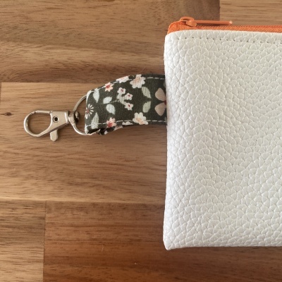 Bolsa branca com fecho laranja, alça floral verde e mosquetão prateado sobre madeira