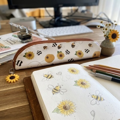 Estojo branco decorado com abelhas e girassóis sobre mesa de madeira com caderno aberto com desenhos da mesma temática.