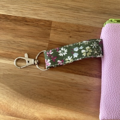 Chaveiro com mosquetão metálico, fita floral verde e bolsa rosa em material sintético.