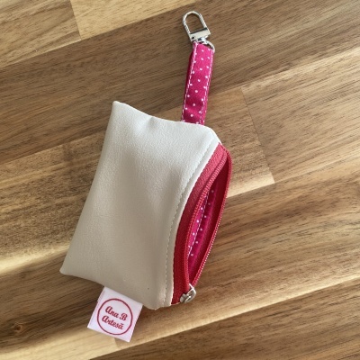 Estojo branco com fecho e pega rosa com pontos brancos, etiqueta Ana.B Artesã sobre mesa de madeira.