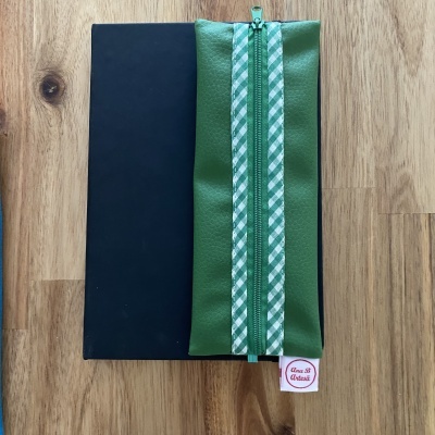 Estojo verde com fecho e padrão xadrez sobre livro preto em madeira clara