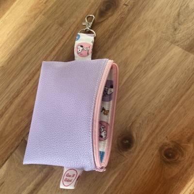 Bolsa lilás texturizada com fecho rosa e asa estampada com personagens, etiqueta Ana B Artesã, em fundo de madeira clara